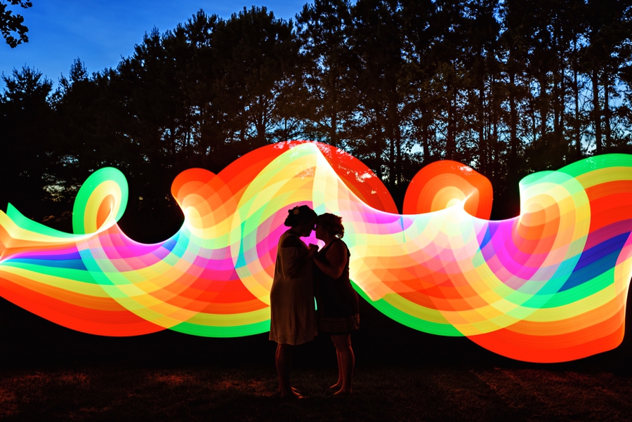 A newly married couple hold each other with LGBTQ+ rainbow swirling behind them.